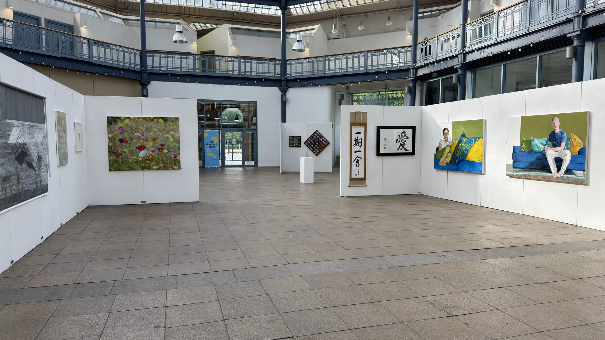 Installation view of the Studio Artists exhibition in the glass-roofed atrium of The Briggait, with two large sofa paintings by Alan Brash on the right-hand wall.
